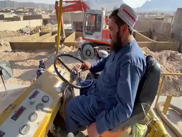 The video shows a man with a beard and a white cap sitting in the driver's seat of a large yellow construction vehicle. He is wearing a blue outfit and holding the steering wheel. Behind him, there is an orange excavator and other construction workers. The background shows a mountainous landscape and a partially constructed building. The scene appears to be a construction site.