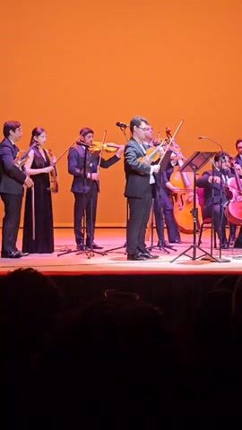 The video captures a live musical performance on a stage. A group of musicians, dressed in formal attire, are playing string instruments. From left to right, there appear to be four violinists, followed by a cellist and possibly another cellist or bassist further to the right. The musicians are positioned on a wooden stage with microphones and stands visible. The backdrop is a gradient of orange transitioning to pink and then purple. The foreground shows the dark silhouettes of an audience watching the performance. The lighting is focused on the stage, creating a dramatic effect.