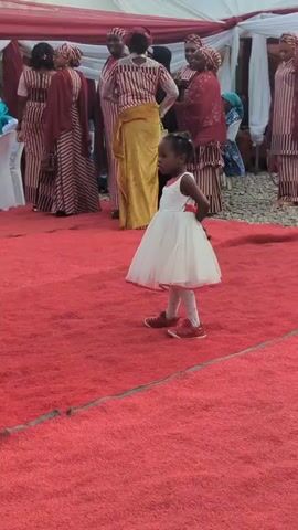 A short video of a young girl with black hair and a white dress standing on a red rug. A person's arm is seen in a gold outfit. The girl is seen on a red rug again and turns around as she stands still. She has red shoes on. Many women are seen behind her in formal attire.