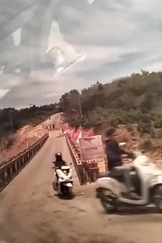 The video shows a first-person perspective from inside a car as it drives across a bridge. The bridge appears to be made of metal and wood with red and white flags on either side. The car is moving forward on a paved road. The surrounding environment consists of hilly terrain with trees. A motorcycle with a rider can be seen on the bridge in the initial frames. The sky is overcast with clouds. A sign is visible on the right side of the road near the bridge entrance. The interior of the car is also visible, including the dashboard, steering wheel, and rearview mirror. Reflections are present on the car's windows, obscuring some of the view. The overall impression is of a scenic drive across a bridge in a rural or mountainous area.