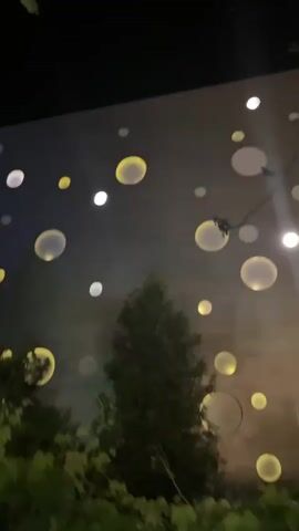 The video shows a nighttime exterior shot of a building with a light-colored facade covered in circular lights of various sizes and colors, some white and some yellow. The building is partially obscured by dark trees and foliage in the foreground. The lights on the building appear to be a decorative lighting installation. The camera pans slightly upwards, revealing more of the building and the dark sky above.
