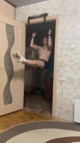 A man is hanging from a pull-up bar installed in a doorway, shirtless and wearing dark underwear and white socks. His legs are straight and held out in front of him. A small girl appears briefly in the left corner of the shot before the pull-up bar dislodges and falls, causing the man to drop to the floor. The doorway has a light-colored frame and a door with a decorative, wavy inlay. The wall is covered in a patterned wallpaper featuring small trees. The room has a striped, round rug on a wooden floor and a white radiator is visible in the background. A small brown cabinet is also visible through the doorway.