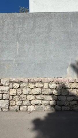A person walks along a stone retaining wall, turns to face the wall, puts their hands on the wall, and jumps off the retaining wall. The person is wearing a light green shirt, dark shorts, gray socks, and Vans sneakers. The retaining wall is made of light gray stones and is in front of a larger gray wall. The sky is blue.