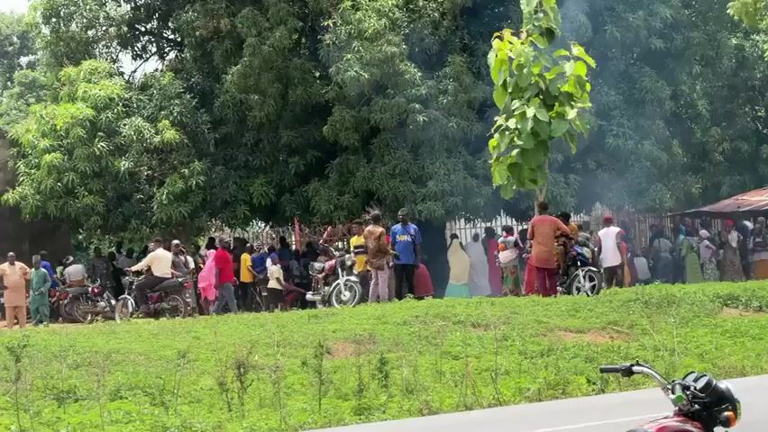 The video shows a group of people gathered outside a building that appears to be a school or community center. There are several motorcycles and vehicles parked nearby. The building is somewhat dilapidated with a damaged roof in places. The surrounding environment is green, with grass and trees. Some people are standing and talking, while others are sitting. The atmosphere appears calm, though the reason for the gathering is unclear. The video is shot from a distance, providing a general overview of the scene.