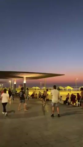 The video captures a group of young adults playing a game, possibly using a small ball or a similar object. The scene takes place on a paved area next to a beach at dusk. The sky transitions from blue to hues of pink and orange. People are sitting or standing on the beach in the background. A modern, flat-roofed structure provides shade and is illuminated with lights. The players are dressed casually, and others are walking by or observing the game. Overall, the scene reflects a relaxed atmosphere during twilight hours.