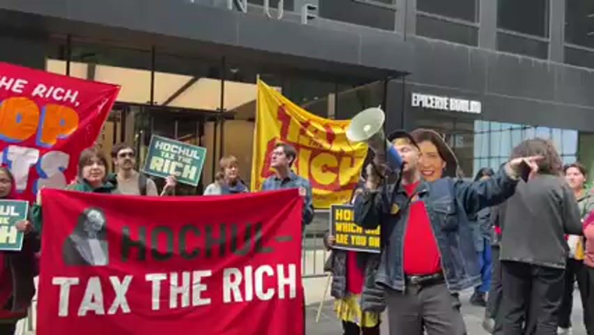 A group of approximately 15-20 people are gathered outdoors on a city street, participating in a protest. The scene is set in front of a modern building with large windows, and the name "EPICERIE BOULUD" is visible on the upper part of the building.

The protesters are holding various signs and banners. In the foreground, a large red banner reads "HOCHUL - TAX THE RICH," featuring a stylized image of a person. Several individuals are holding smaller green signs with the same message, "HOCHUL TAX THE RICH." A yellow banner in the background also displays "TAX THE RICH" and possibly other text like "STOP THE CUTS, FUND OUR FUTURE." Another red banner on the far left has "THE RICH, STOP THE CUTS" partially visible. A person near the megaphone holds a sign asking, "HOCHUL WHICH SIDE ARE YOU ON?"

A man in the center of the frame is speaking into a megaphone. He is wearing a blue denim jacket over a red shirt, and he has a large cardboard cutout of a woman's head (resembling New York Governor Kathy Hochul) attached to his own head. The protesters are actively chanting in unison, repeatedly saying "Tax the rich!" followed by "Stop the cuts!" The overall atmosphere is one of organized protest and advocacy. The weather appears to be clear and bright, indicating daytime.