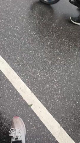 The video shows the perspective of someone standing on a wet asphalt surface, likely a road or parking lot, as indicated by the grey, speckled texture and a white painted line. Several pairs of feet and legs are visible, suggesting the presence of multiple individuals. In the foreground, on the left, is a pair of white and grey athletic shoes with pink accents and navy socks. To the right, partially in motion, is a person wearing black boots. Further to the right, and also moving, are legs clad in dark grey pants, with black and yellow athletic shoes. Another pair of legs, wearing dark trousers and black shoes with white soles, is visible at the top of the frame. The ground is wet and reflective, with small puddles visible. A single green leaf rests on the white line. The overall impression is of people standing or walking in damp conditions.