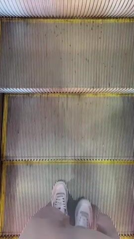 The video shows an upward-facing shot of an escalator. The camera is positioned at the bottom of the escalator, looking up as it moves. The steps are metallic with ridges. The yellow safety lines are visible on the edges of the steps. The feet of a person wearing white sneakers and a gray skirt are visible at the very bottom of the frame, standing on the escalator. The escalator is moving upwards, indicated by the changing perspective and the appearance of more steps as the video progresses.