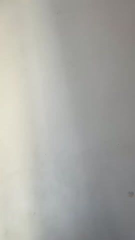 The video shows a white wall with an air conditioning unit mounted high up on it. The camera is angled upwards towards the air conditioner, and the wall appears to be illuminated by a light source from the left, casting a shadow towards the right. The air conditioner has black slats and a white casing. There are a few faint marks on the wall.