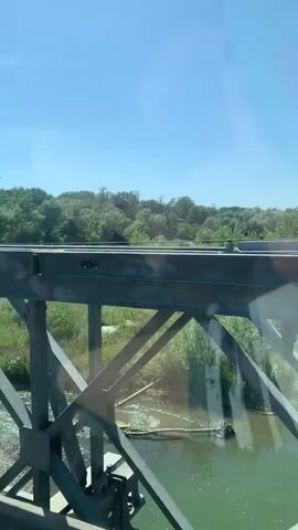 This video shows the view from inside a car as it drives across a bridge. The camera is positioned to show the dashboard, the legs of a passenger, and the view out the side window. The bridge appears to be made of metal and wood, and the car is moving at a moderate speed. The surrounding environment is green and lush, with trees and vegetation visible on either side of the river below the bridge. The sky is clear and blue.
