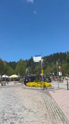 The video shows a sunny day with a clear blue sky and some clouds. It appears to be a town square or a busy street in a European town, possibly in Germany, given the street signs that are visible. There is a circular island in the center of the road with a large display of yellow flowers. Several cars are parked and driving around the roundabout. There are also shops and outdoor cafes with people sitting and walking around. Trees and a forest are visible in the background. The camera pans and moves to show different angles of the scene. One shot focuses on a street signpost with multiple directional arrows. Another shot shows people walking along a sidewalk lined with shops and umbrellas. The overall impression is one of a lively and pleasant summer day in a town.