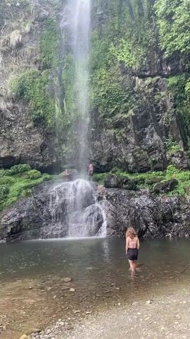 The video depicts a natural scene featuring a waterfall cascading into a small pool. The waterfall is surrounded by lush, green vegetation growing on rocky cliffs. In the foreground, a woman stands in the shallow water of the pool, facing towards the waterfall. Further towards the waterfall, two men can be seen standing on the rocks at the base of the falls. The water appears clear and the environment seems tranquil and natural.