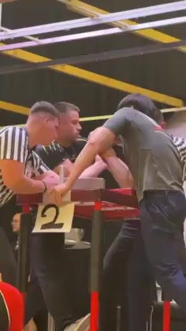 A scene capturing an arm wrestling match in progress. Two men are intensely engaged in the competition at a table marked with the number "2". A referee, clad in a black and white striped shirt, watches closely, ready to intervene as needed. Another man, possibly another referee, stands to the right. The environment suggests an organized arm wrestling event, with overhead lighting and additional individuals in the periphery observing the match.