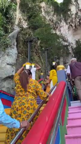 The video shows a group of people ascending a brightly colored staircase. Several of them, mostly women, are carrying what appear to be silver pots on their heads, adorned with yellow flowers. The staircase is painted in vibrant colors and has metal handrails. The setting appears to be outdoors, with large rock formations and trees forming the backdrop. The lighting suggests it's daytime. The people are dressed in casual clothing, with some wearing traditional attire. They are moving upwards, presumably towards a destination at the top of the stairs.