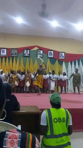 Here's a concise description of the video:

The video shows a cultural performance taking place indoors, likely in a community hall or auditorium. On a red-carpeted stage, a group of people, mostly women, are dressed in traditional or themed outfits. The men stand out in their outfits.

One man in particular, center stage, is wearing a loincloth-style garment with African prints and is carrying something that appears to be a white horn or tusk. The background is decorated with red and gold curtains, and framed pictures are hanging above them.

The audience includes people wearing uniforms, possibly security or event staff. One person in the foreground is wearing a bright green vest labeled "OBS". The overall atmosphere suggests a formal event or cultural celebration.