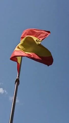 A Spanish flag flies against a blue sky with scattered white clouds. The flag is attached to a tall metal pole and is shown waving in the wind. The flag has horizontal stripes of red and yellow, with the coat of arms of Spain on the yellow stripe. The video is filmed from a low angle, looking up at the flag and sky. The sun is visible, casting a bright light on the flag. There are no people or other objects visible in the scene. The video is approximately 5 seconds long.