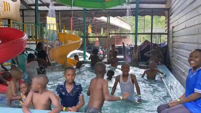 The video shows a group of children, mostly shirtless, enjoying themselves in a brightly colored indoor water park. The pool is surrounded by playground equipment, including a red slide and a yellow spiral slide. Water is cascading down from overhead structures, adding to the fun. Adults can be seen supervising the children. The atmosphere appears lively and joyful, with the kids splashing and playing in the water. The park is enclosed by walls with window openings revealing the outdoors.
