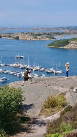 The video shows a scenic view of a harbor with numerous sailboats docked. On a rocky outcrop overlooking the harbor, two people are visible. One person, on the left, is wearing a patterned dress and appears to be taking a photograph with a phone. The other person, on the right, is wearing a yellow top and blue jeans and is also holding up a phone, likely taking a picture or recording. The landscape includes islands in the distance and vegetation on the right side of the frame. The weather appears sunny and clear.