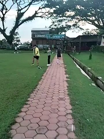 The video depicts an outdoor scene of people playing football (soccer) on a green grass field, likely in Matesih, Indonesia. Several individuals, predominantly men, are actively engaged in the game, running, passing, and kicking a football. Some players are seen wearing jerseys, while others are in casual athletic attire.

At the start of the video, two people are visible on a paved pathway made of hexagonal bricks alongside the field. One individual is performing what appears to be warm-up exercises or stretching. The camera then pans to show more of the field and the ongoing game.

The setting includes large, mature trees with dense canopies and several palm trees, suggesting a tropical or subtropical environment. In the background, beyond the trees, some buildings are visible. The sky is overcast and cloudy, casting a soft, diffused light over the scene, indicating it is likely late afternoon or early evening. The overall impression is of a casual or recreational football session taking place in a community or local sports ground. No discernible text is visible in the video.