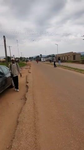 A male runner in a pink tank top and black shorts runs towards the camera along a paved road. He is followed by a white van with flashing lights, several motorcycles, and a group of people. A man standing on the side of the road watches and films the runner. Other people stand by the road, including one person taking pictures. The road is lined with buildings and trees, and the sky is overcast.