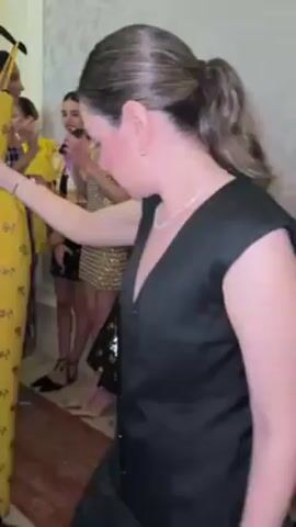 The video shows a rack of clothing. A yellow dress with a rose print hangs at the center. To the side is a white dress with black polka dots, and a red rose floral print top and purple shorts hanging next to that. There are images of women modeling the clothing pinned above each garment.  A woman in a black v-neck top and pants is visible moving around the rack of clothes. Other individuals are visible in the background.