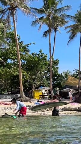Two people carry surfboards from the water onto a beach on a sunny day. One person is wearing a light blue shirt and red shorts, while the other is wearing dark shorts. They carry a surfboard each. The water is clear and shallow near the shore. The beach is sandy with palm trees and other tropical vegetation. Other people are visible on the beach relaxing and walking around. There is a small yellow trailer-like structure and sun shelters visible inland from the shore. The sky is clear and blue.