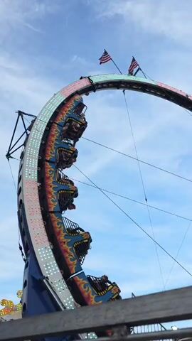 This media file captures a thrilling amusement park ride, likely a pendulum or looping ride, in action. The ride features a large, curved structure with rows of seats, some of which are occupied by riders who appear to be experiencing the excitement of the ride. The ride itself is decorated with vibrant colors and patterns, including flames, and is adorned with numerous lights. Two American flags are prominently displayed at the apex of the ride's arc.

In the background, the sky is a clear blue with wispy clouds, suggesting a pleasant day, possibly late morning or afternoon. Another amusement ride, featuring a sign that reads "FIRE BALL," is visible to the right, indicating a carnival or fair setting. The foreground shows a railing or fence, suggesting the viewer is observing the ride from a safe distance. The overall atmosphere appears lively and energetic, typical of a public event or fairground. The image suggests a sunny day at a place of entertainment.
