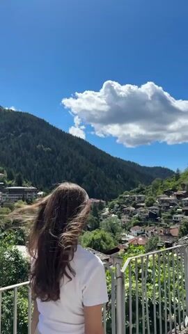 A young woman with long, dark brown hair stands with her back to the camera, looking out over a scenic village nestled in a valley. The village is comprised of numerous houses with red-tiled roofs, clustered on the hillside. Lush green trees and a dense forest cover the slopes of the surrounding mountains. The sky is a vibrant blue, with a large, fluffy white cloud dominating the upper portion of the frame. The woman is wearing a white t-shirt and a light-colored skirt. She is standing on a balcony or observation deck, with a white railing in the foreground. The scene is bathed in bright sunlight, indicating a clear, sunny day.