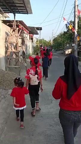 In a residential street, a group of children, mostly dressed in red shirts and some in black pants, are walking in a line. Two young girls in the foreground are holding hands, both wearing red tops and black leggings. The girl on the left has her hair in pigtails, while the girl on the right wears a red cap and has a backpack on. Behind them, a person wearing a black headscarf and a red top is walking away from the camera. The street is paved and appears to be a narrow alleyway lined with buildings and trees. There are power lines overhead, and flags are visible in the distance. The lighting suggests it is daytime, possibly late afternoon.