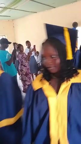 The video shows a group of graduating students marching through a room. They are wearing blue graduation gowns with yellow accents and graduation caps. Several people are standing on the sidelines, watching and taking pictures with their phones. The room has white walls, a light green ceiling, and white plastic chairs lined up against one wall. There are balloons in the corner of the room. Music is playing in the background. The video seems to be shot from the perspective of someone moving with the graduating students.