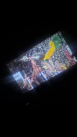 The video displays a screen in a dark room, showing a sports celebration. The initial scene features multiple people holding up a trophy amidst falling confetti. A green and yellow flag is visible in the background, along with a partial word "ADORES" on a sign. The scene suggests a championship victory, possibly in football.

The video then transitions to a list of years displayed vertically on the screen: 2008, 2009, 2010, 2011 (highlighted in red), 2012, 2013, 2014, and 2015. This suggests a timeline or a retrospective of achievements. The screen has a bright reflection on its surface. The overall setting is a person watching content on a screen in a dimly lit environment.