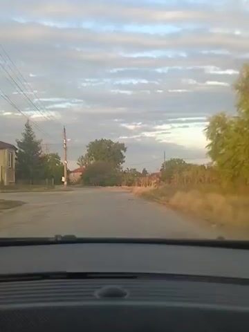 The video shows a view from inside a vehicle, looking out through the windshield at a road in a rural area. The sky is partly cloudy with hints of sunset. Trees and houses are visible on either side of the road. A power pole with wires is on the left. Another vehicle is seen driving in the distance on the road. The road appears to be paved but has some cracks and unevenness. The overall impression is of a calm, possibly early evening scene in a village.