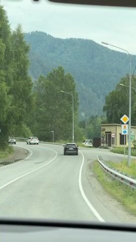 This is a first-person view from inside a vehicle, looking out at a road in a rural, mountainous area. The road curves to the left, with a white car and a black SUV in front of the camera. To the right of the road, there is a small yellow building with a sign that reads "МАГАЗИН" (Magazin), indicating it is a shop. Next to the shop, there are several signs, including a diamond-shaped yellow sign with a black border and a red circle with a line through it, which is a traffic sign. The shop has display windows and appears to be open.

Behind the shop and the road, a dense forest covers the rolling hills and mountains. The trees are lush and green, suggesting it is summertime. The sky is overcast, with diffused light, indicating either early morning, late afternoon, or a cloudy day. There are streetlights visible along the road, and a utility pole stands near the shop. The overall scene conveys a sense of travel and a quiet, scenic environment. The presence of the shop suggests a small settlement or a stop along a route. The visible text "МАГАЗИН" is the only legible Russian text in the frame. The weather appears to be mild and possibly damp, given the overcast sky. The mood is neutral, focusing on the journey and the surrounding landscape.