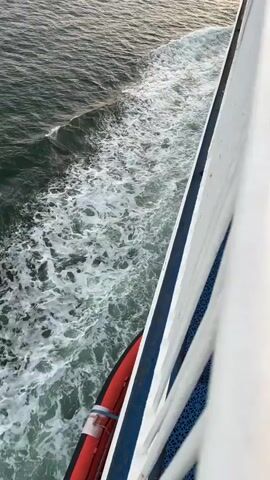 The video shows the wake of a large vessel moving through the ocean. The camera is positioned on the deck of the vessel, looking out towards the water. The water is choppy, with white foam churned up by the boat's passage. On the right side of the frame, a white railing with blue decking is visible. Towards the back of the frame, a life raft is mounted on the side of the boat. The sky above is a soft orange and yellow, suggesting either sunrise or sunset. Distant ships can be seen on the horizon.