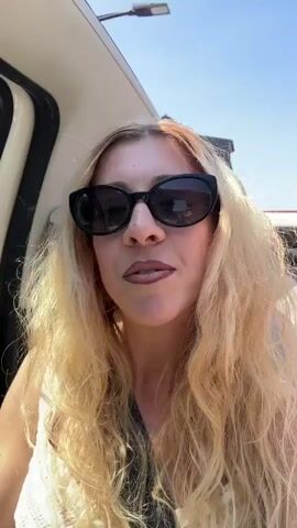 A blonde woman wearing sunglasses and a white top is in the passenger seat of a car. She is looking at the camera and speaking. Her mouth is open and she is animated. The car appears to be moving. The background shows a cobblestone street, a pink building, trees, and a clear blue sky.
