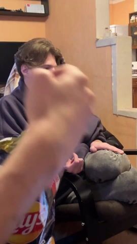 The video shows two men in an indoor setting. One man, with brown hair, a beard, and wearing a purple hoodie, is seated in a black chair. Someone hands him a glass bottle. The second man, wearing glasses and a black t-shirt and pants, is kneeling on a pillow on the floor. A laptop, a mouse, and some cables are visible near him on the floor. The wall has an orange floral wallpaper.