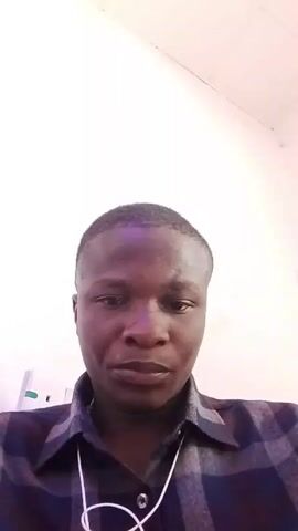 A close-up video shows a dark-skinned man with a shaved head wearing a plaid shirt and a white wristwatch. He looks up at the camera and puts his hand to his chin, appearing thoughtful. The background is a plain white wall.