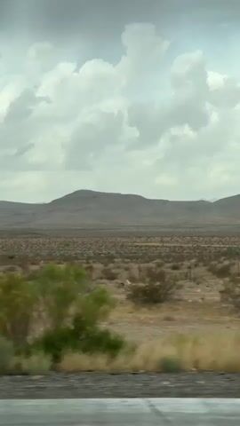 The video displays a desert landscape under a cloudy sky. A large billboard is visible in the distance, standing tall on a metal pole. The billboard has two sections, one on the left with colorful imagery and text, and another on the right with text that is partially obscured. The surrounding environment is barren, with sparse vegetation and rolling hills in the background. The camera appears to be moving, likely from a vehicle, as evidenced by the blurred foreground elements and the sense of passing scenery. The overall impression is one of a vast, arid expanse with a solitary man-made structure.