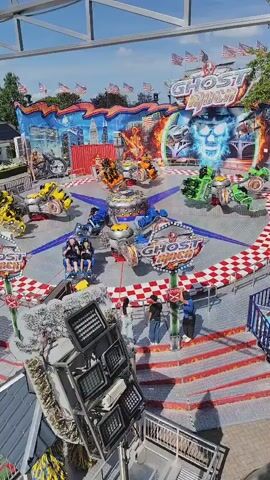 This is a high-angle, medium shot of a carnival ride called "Ghost Rider" on a sunny day. The ride features multiple motorcycle-themed vehicles that spin and move in a circular pattern around a central hub. The background of the ride is decorated with imagery of a skull with glowing blue eyes and flames, along with urban cityscapes and American flags. Several people are visible standing around the ride, observing or waiting to ride. One person is in the foreground, standing on a platform with a large, metallic structure with multiple lights attached to it. The ground around the ride is covered in red and white checkered flooring, and there are red steps leading up to the ride platform. The overall environment appears to be a bustling outdoor amusement park or fair.
