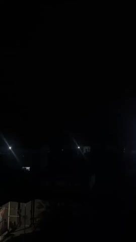 The video shows a nighttime scene. Initially, the sky is completely dark with a few distant lights visible. Around the 3-second mark, a flash of lightning illuminates the sky, revealing dark clouds and silhouetted buildings below. The scene then returns to darkness.
