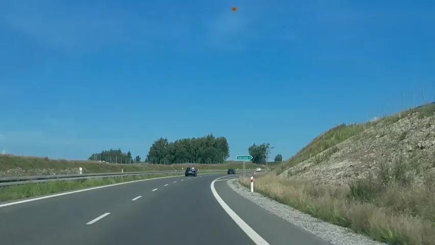 The video shows a view from a vehicle driving on a highway under a clear blue sky. The highway has multiple lanes, indicated by white lines. Several cars are visible on the road. On the right side of the highway, there is a grassy embankment with some rocks and a few white and red marker posts. On the left side, there is a barrier and a line of trees. A green road sign with Polish text and the number 5 is visible in the distance. The camera appears to be mounted on the dashboard or windshield of the vehicle, providing a forward-facing perspective of the road ahead.