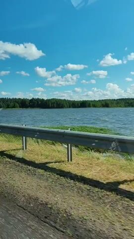 A side view from a vehicle traveling alongside a body of water on a sunny day. A metal guardrail runs along the edge of the road, separating it from the grassy embankment and the lake. Trees line the far shore of the lake, and white clouds dot the bright blue sky. The road surface in the foreground appears to be dirt and gravel.