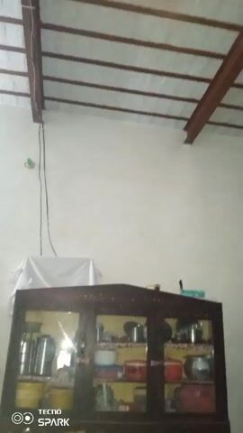A video pans across a room, seemingly a bedroom or living area in a Pakistani home. The ceiling has exposed beams and rafters. A large wooden cabinet with glass doors is filled with various kitchen items, including pots, pans, and containers. To the left of the cabinet, a white sheet is draped over something. Further to the left, a light fixture hangs from the ceiling. A small green object is visible on the wall. In the center of the room, a reddish-brown cabinet or chest of drawers is present. To the right, a shelf unit is laden with numerous items, including jars, boxes, and possibly food items. A red refrigerator is positioned to the left of the shelf unit. A bed with a patterned comforter is visible in the foreground. The overall impression is of a functional, lived-in space.