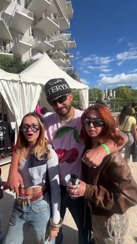 A man and two women pose for a photo outdoors. The man is in the center, wearing a pink shirt with cherries, a black beanie with the word "ARCTERYX" on it, and sunglasses. He has his arms around the shoulders of the two women. The woman on the left has long blond hair and is wearing a gray top, blue jeans, and sunglasses. The woman on the right has reddish-brown hair and is wearing a brown leather jacket, patterned pants, and sunglasses. All three are holding cups with visible logos. Behind them are white tents, trees, and a tall white building with several balconies. The sky is blue with some clouds. There are other people walking around in the background.