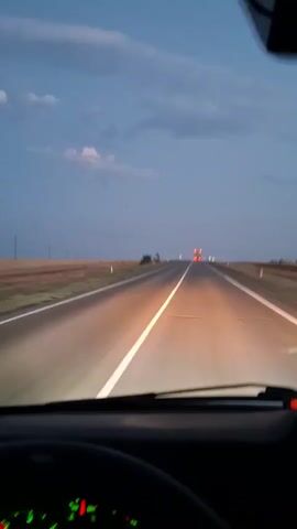The video shows a first-person view from the driver's seat of a vehicle traveling on a highway at dusk. The headlights of the vehicle illuminate the road ahead, with a white line marking the center. In the distance, the taillights of other vehicles are visible. The sky is a deep blue with scattered clouds, and the landscape on either side of the road appears to be open fields. The dashboard of the vehicle is visible in the foreground, with illuminated gauges and a digital display. The video appears to be shot from a moving vehicle.