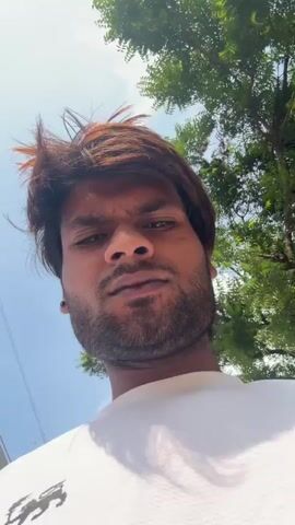 A man with brown hair and a beard looks up into the camera. He is wearing a white t-shirt. The background consists of green trees and a bright blue sky with some clouds. He raises his hand to his head and runs his fingers through his hair.