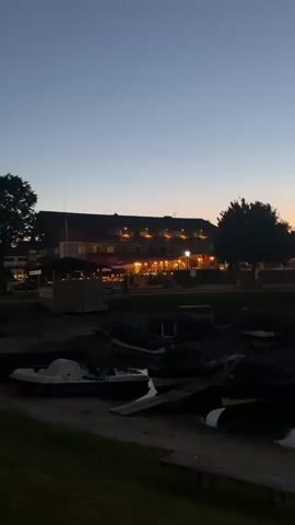 The video shows a still shot of a lake at dusk. Boats are docked on the shore, covered with tarps. In the background, a building with lights on is visible, along with trees and tents. The sky is a gradient of blue and orange, indicating sunset. The overall scene is tranquil and serene.