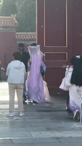 A group of four women in traditional Chinese dresses are walking away from the camera on a stone-paved path. Three of the women have long, dark hair, and one is wearing a long, lilac-colored dress with a veil. Two people in casual clothing follow behind them. The group is passing through a large, red doorframe with a blue sign above. The surroundings suggest a historical or cultural site.