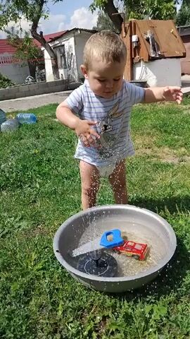 A toddler wearing a diaper and a striped t-shirt plays with toys in a metal basin of water. The child is splashing water with their hands. In the basin are a blue toy truck, a red toy car, and a solar-powered fountain. The scene takes place outdoors on a sunny day, with grass and a building in the background.
