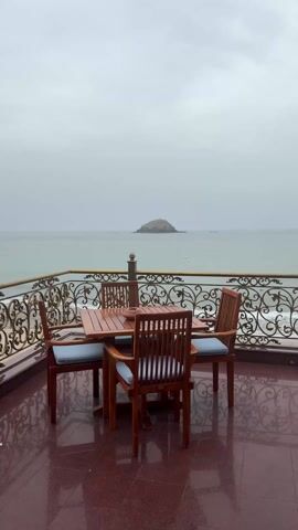 The camera slowly approaches a patio table and chairs, crafted from dark wood with light blue cushions. The patio has a dark red-brown tile floor with a repeating pattern. The railing is ornate wrought iron. Beyond the railing is the sea, with a small rocky island in the distance under a grey, overcast sky. The sea is calm, with gentle waves breaking on a sandy beach.
